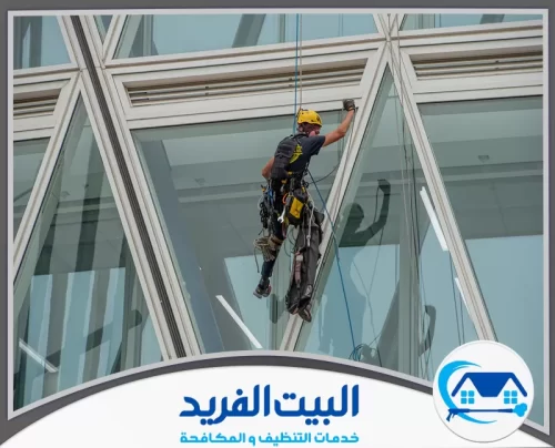 شركة تنظيف واجهات زجاجية مجربة في الشارقة 0567627005 شركة البيت الفريد عامل صيانة محترف يرتدي خوذة ومعدات تسلق كاملة، يتدلى بالحبال لتنظيف أو إصلاح واجهة زجاجية ضخمة لبرج عصري